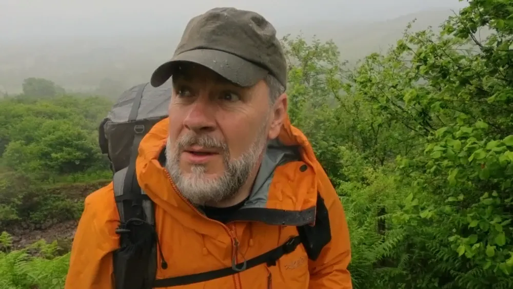 Jim in an orange jacket talking to the camera on a green hillside