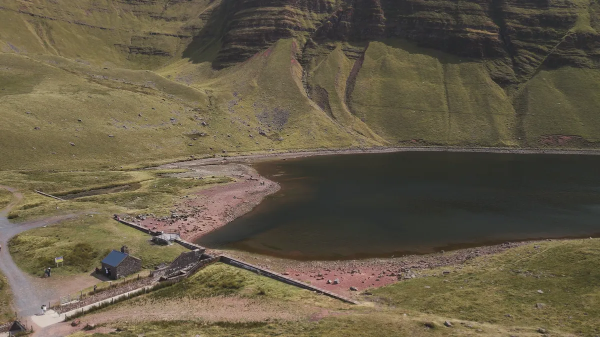 The stone dam and small hut at Llyn y Fan Fach from above, low water exposing the red shoreline below the escarpment