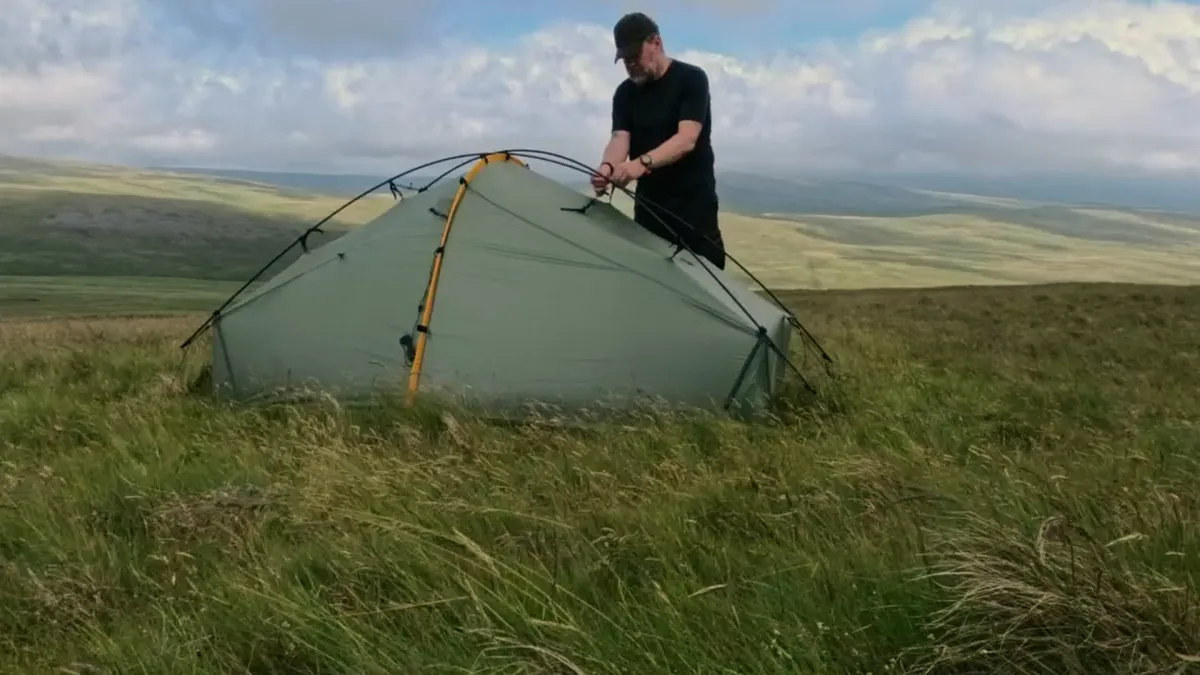 Pitching a green tent on open grassy hillside under a building sky