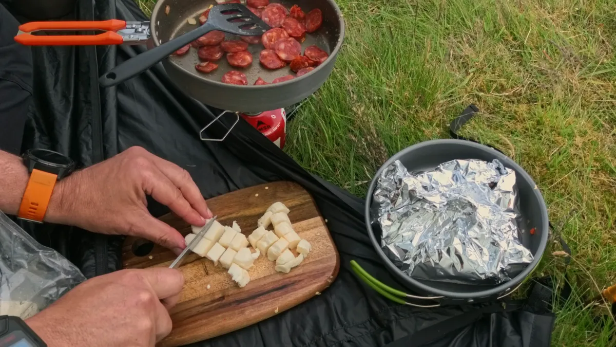 Cubing halloumi on a wooden board while sliced chorizo fries in the pan behind, the potatoes and veg wrapped in foil to one side