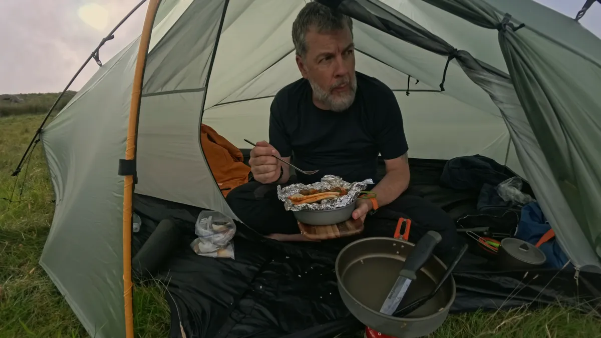 Jim sitting in the doorway of his tent on the open hillside, eating the hash from a foil parcel