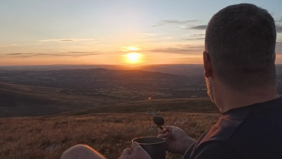 Jim from behind, eating from a titanium mug while watching the sun set over the lowlands far below