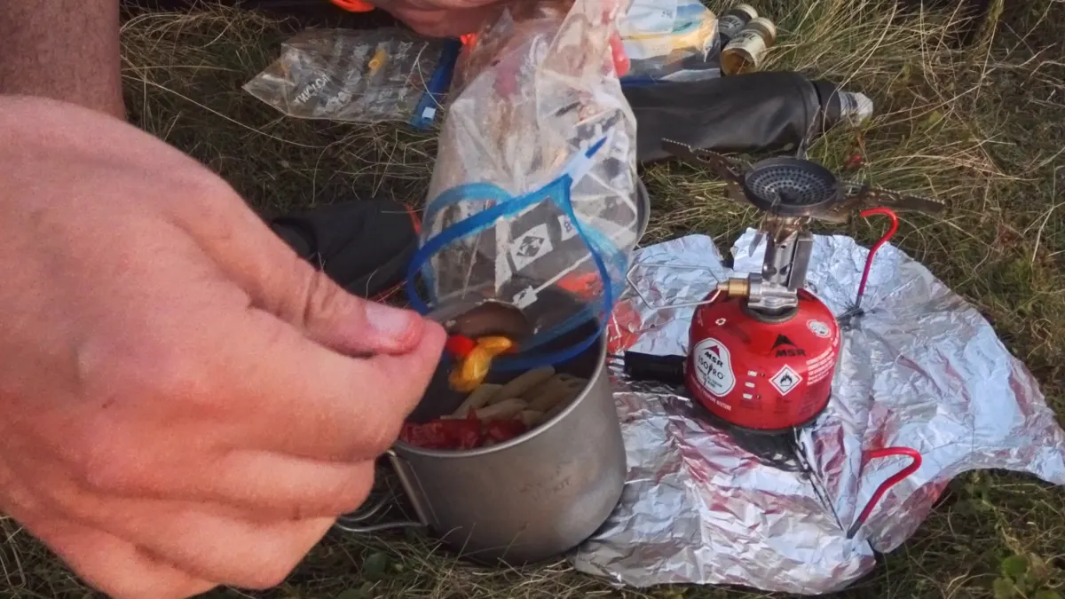 Adding sun-dried tomatoes and roasted peppers from a zip bag to a titanium pot, a small gas stove on foil beside it
