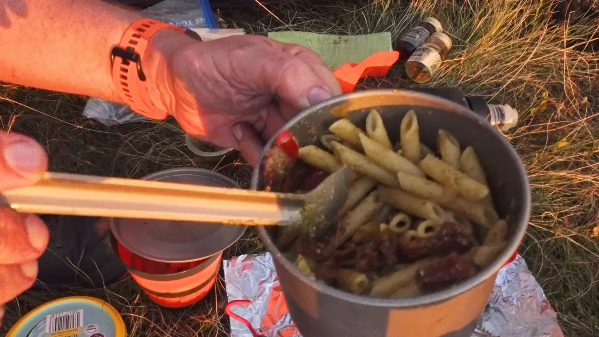 Penne with pesto and sun-dried tomatoes being stirred in a titanium pot on the grass, an orange collapsible cup beside it