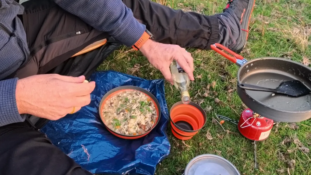 A bowl of mushroom risotto on a blue groundsheet while the last of a small bottle of white wine is poured into an orange collapsible cup