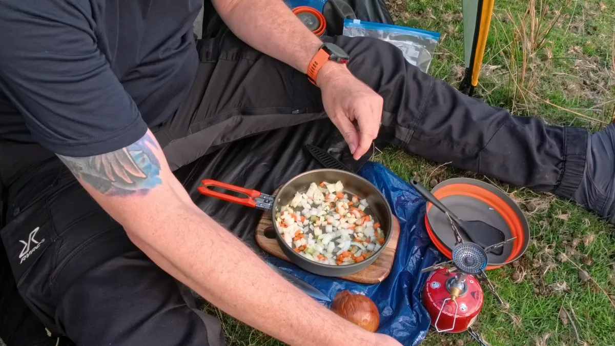 Chopped onion, carrot and pepper frying in a pan on a small gas stove, a titanium pot of stock beside it on a blue groundsheet