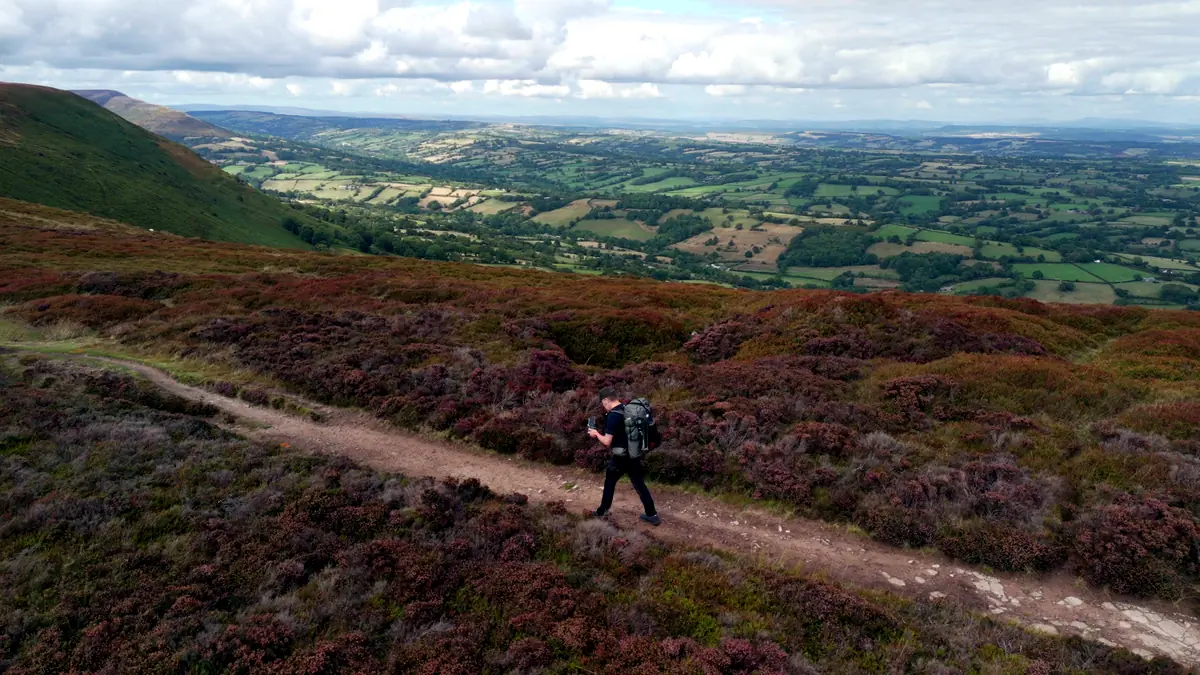 A walker with a pack on a path through purple heather on Hatterrall Ridge, farmland spreading out below