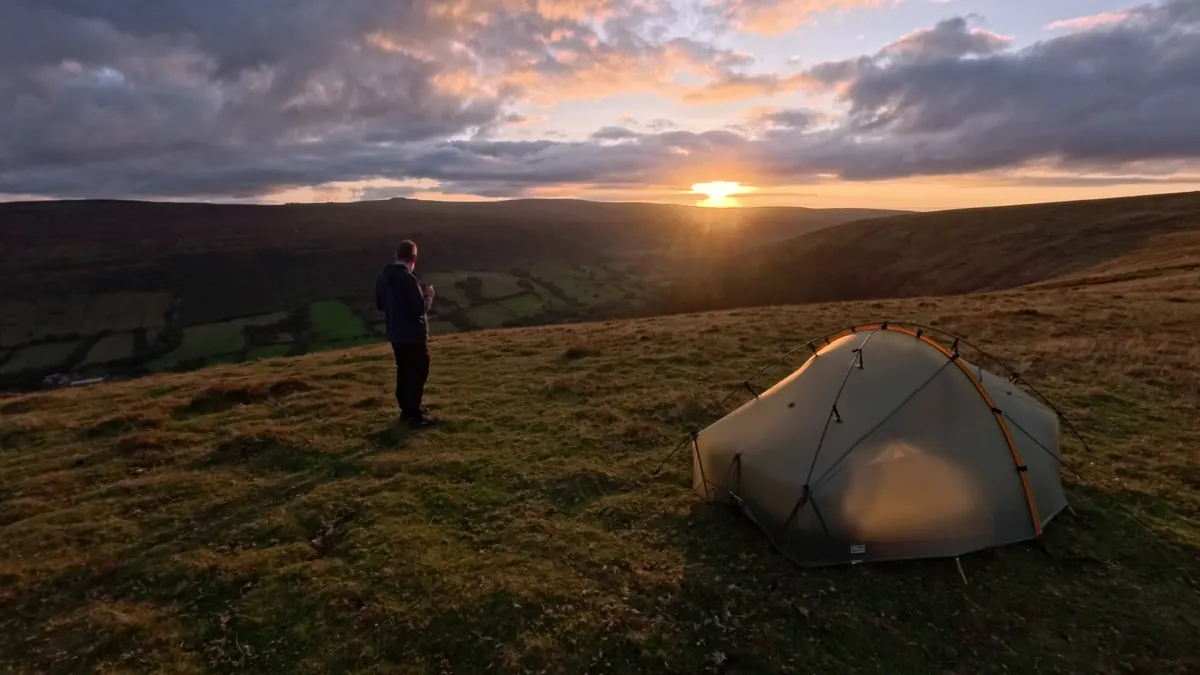 A walker standing on the hillside at sunset beside a small tent lit by the low sun