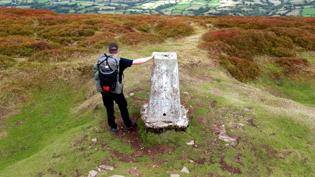 A walker resting a hand on a white trig pillar on the heather ridge